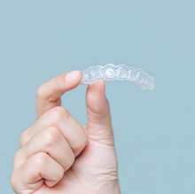 A hand holding up an Invisalign tray with 2 fingers with pale aqua background